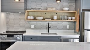 A modern kitchen with a stainless steel refrigerator, double sink, and built-in oven. There are wooden shelves above the sink holding dishes and glasses.