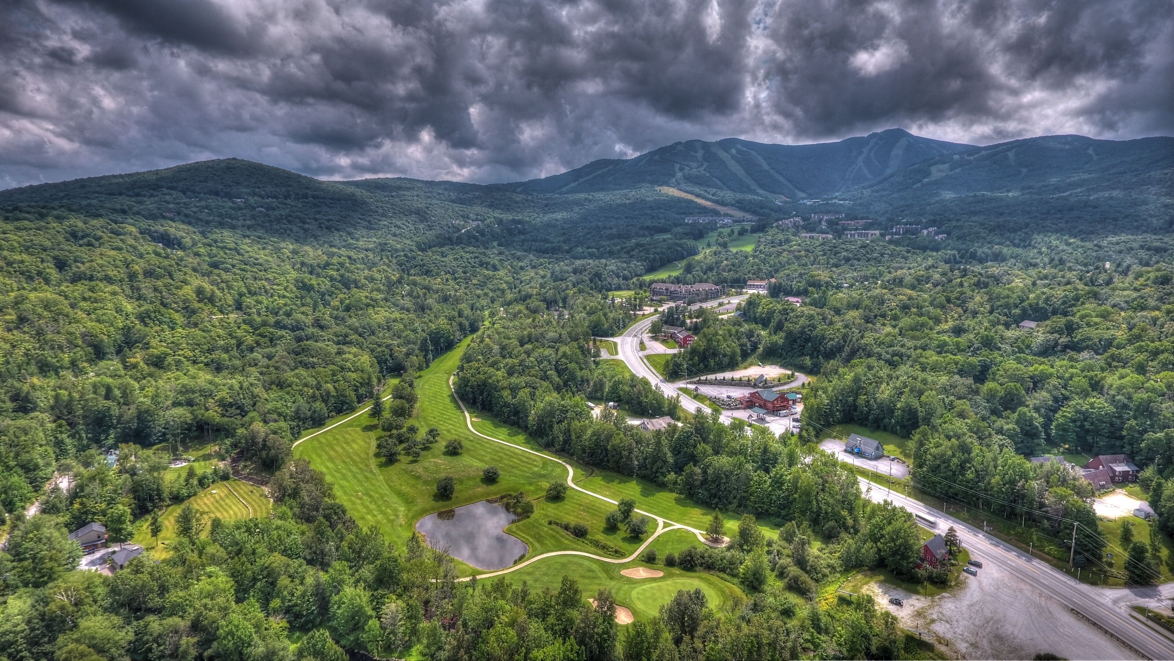 A golf course with a clubhouse, a pond, and a road leading to a residential area surrounded by mountains.