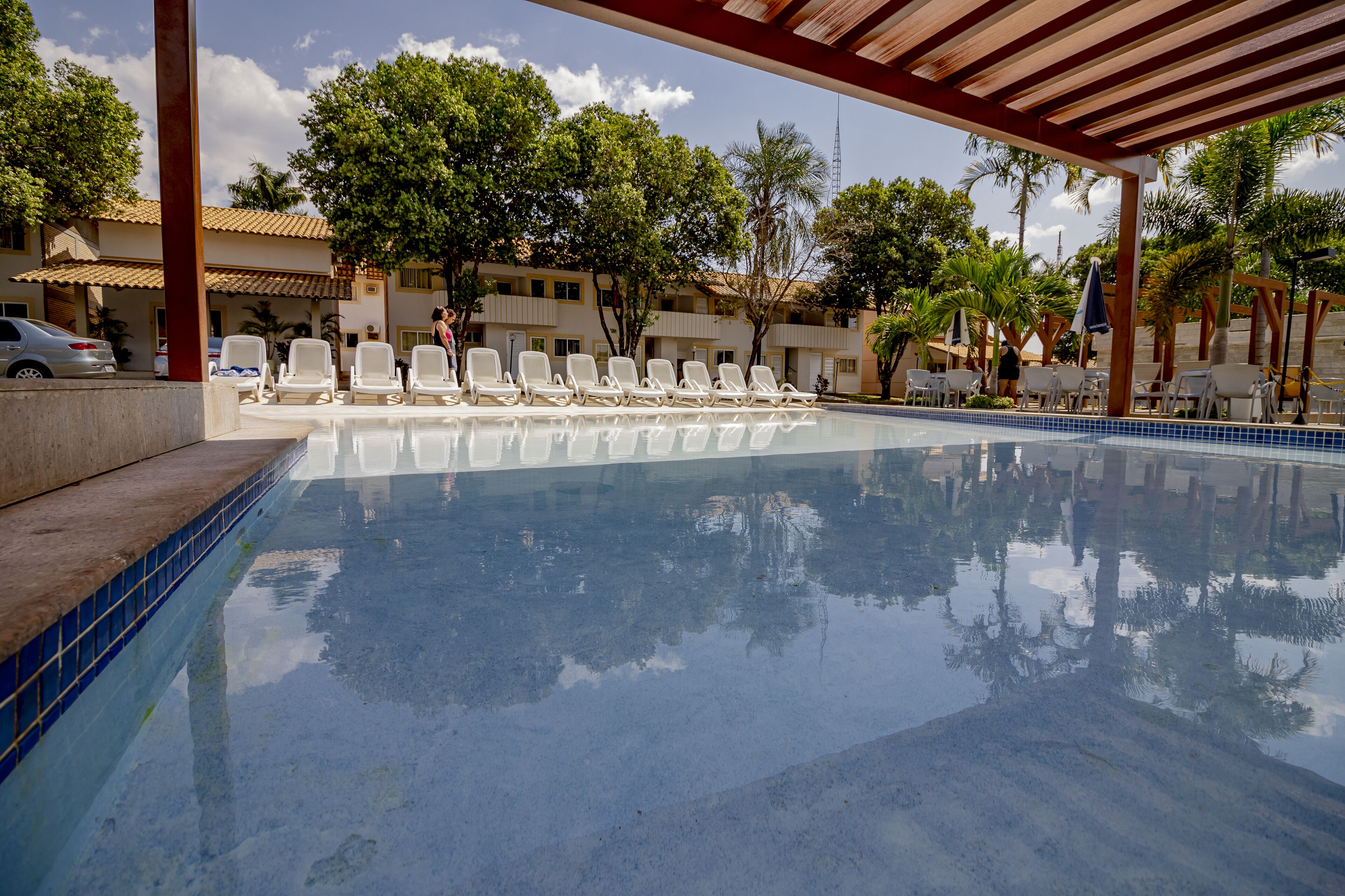 Piscina com espreguiçadeiras e área coberta, cercada por árvores e edifícios.