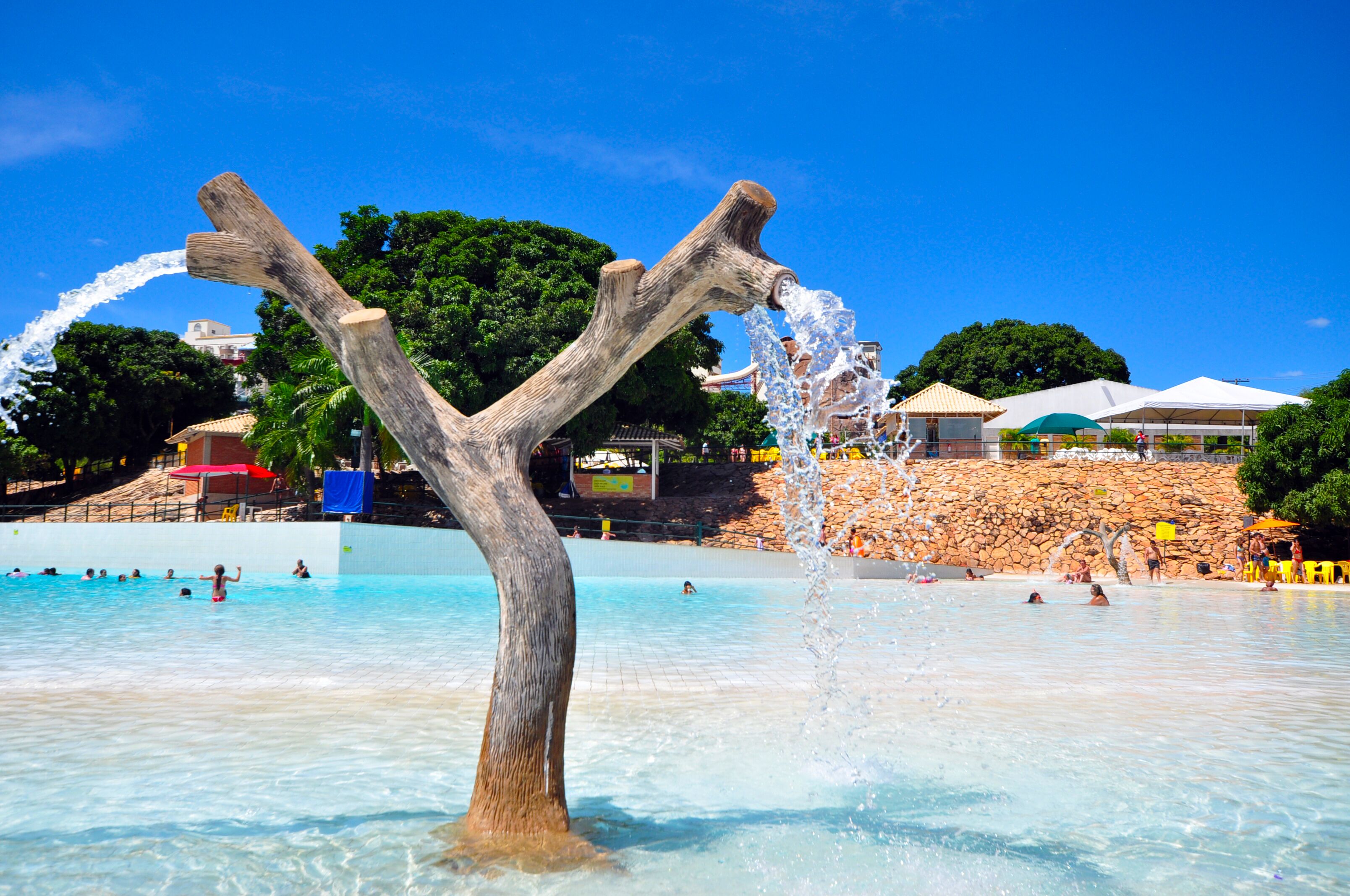 A water feature with a wooden structure in a pool area, surrounded by people and a stone wall with umbrellas.