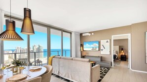Una sala de estar moderna con sofá, mesa de comedor y lámparas colgantes. Un televisor está montado en la pared y se aprecia vista de la ciudad a través de amplios ventanales.