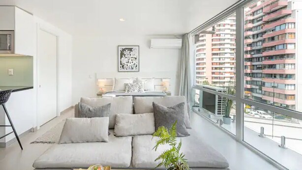 A modern living room with a large sofa, a dining table, and a view of a high-rise building. - New apartment in Barrio El Golf (Las Condes)