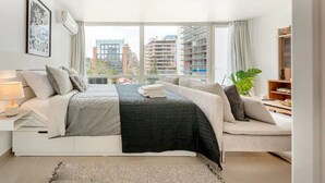A modern bedroom with a large bed, a sofa, and a view of city buildings. - New apartment in Barrio El Golf (Las Condes)