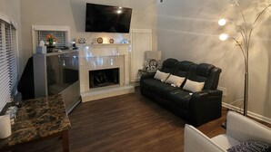 A living room with a fireplace, a black leather sofa, a white armchair, a marble coffee table, and a flat-screen TV mounted on the wall. - Luxury Home 12 Overnight Guests, Wi-Fi for Online Worker, Theme Gold & Diamonds (Woodstock)