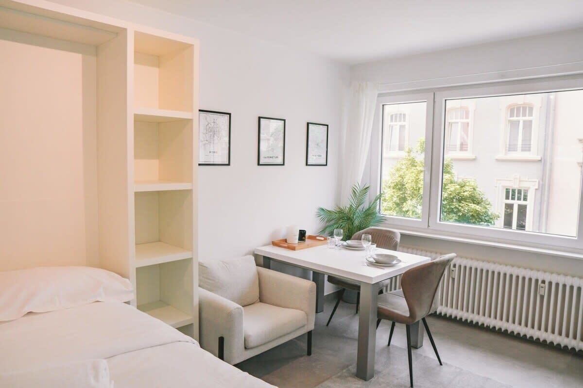 Une pièce moderne et bien éclairée, avec un lit, une table à manger et des chaises, ainsi qu’une fenêtre donnant sur des immeubles. Des étagères et des tableaux encadrés ornent le mur.