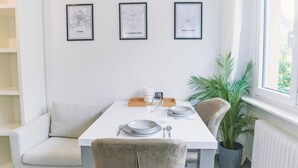A dining area with a white table set for two, framed city maps on the wall, and a potted plant by the window.