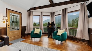 Habitación con pisos de madera, dos sillones verdes, una puerta de madera y ventanas amplias con vista a la montaña.