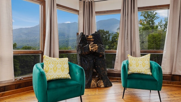 Una habitación con dos sillones verdes, una escultura de madera y ventanas amplias con vistas a la montaña.