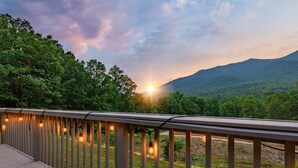 Una terraza de madera con luces colgantes, con vistas a un paisaje boscoso y una montaña al fondo.
