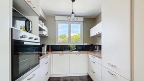 A compact kitchen with white cabinets, a built-in oven, a window with a view, and a hanging light fixture. - Charming house on the heights of St Jacques with sea view (Sarzeau)