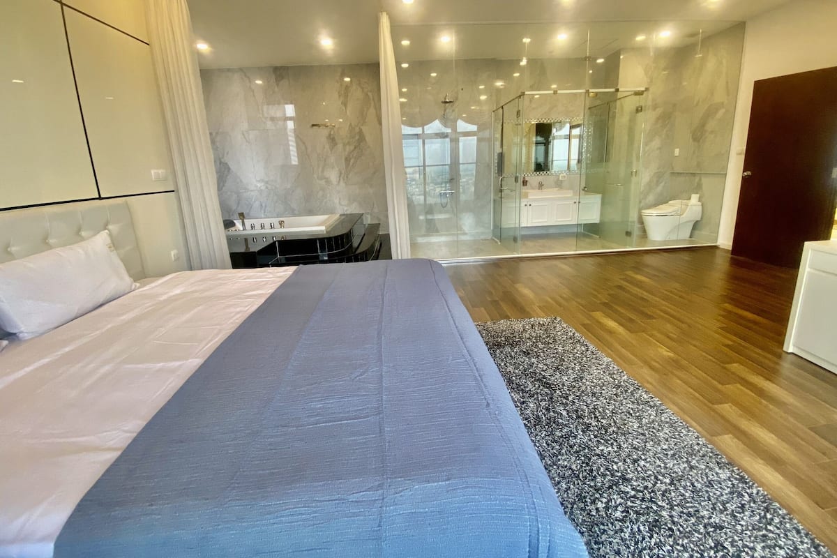 A modern bedroom with a large bed, a bathroom with a glass shower, and a wooden floor.