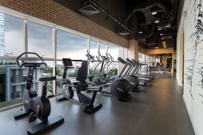 Une salle de sport équipée de tapis de course, de vélos d’appartement et d’appareils elliptiques, dotée de grandes fenêtres et d’un mur en briques.