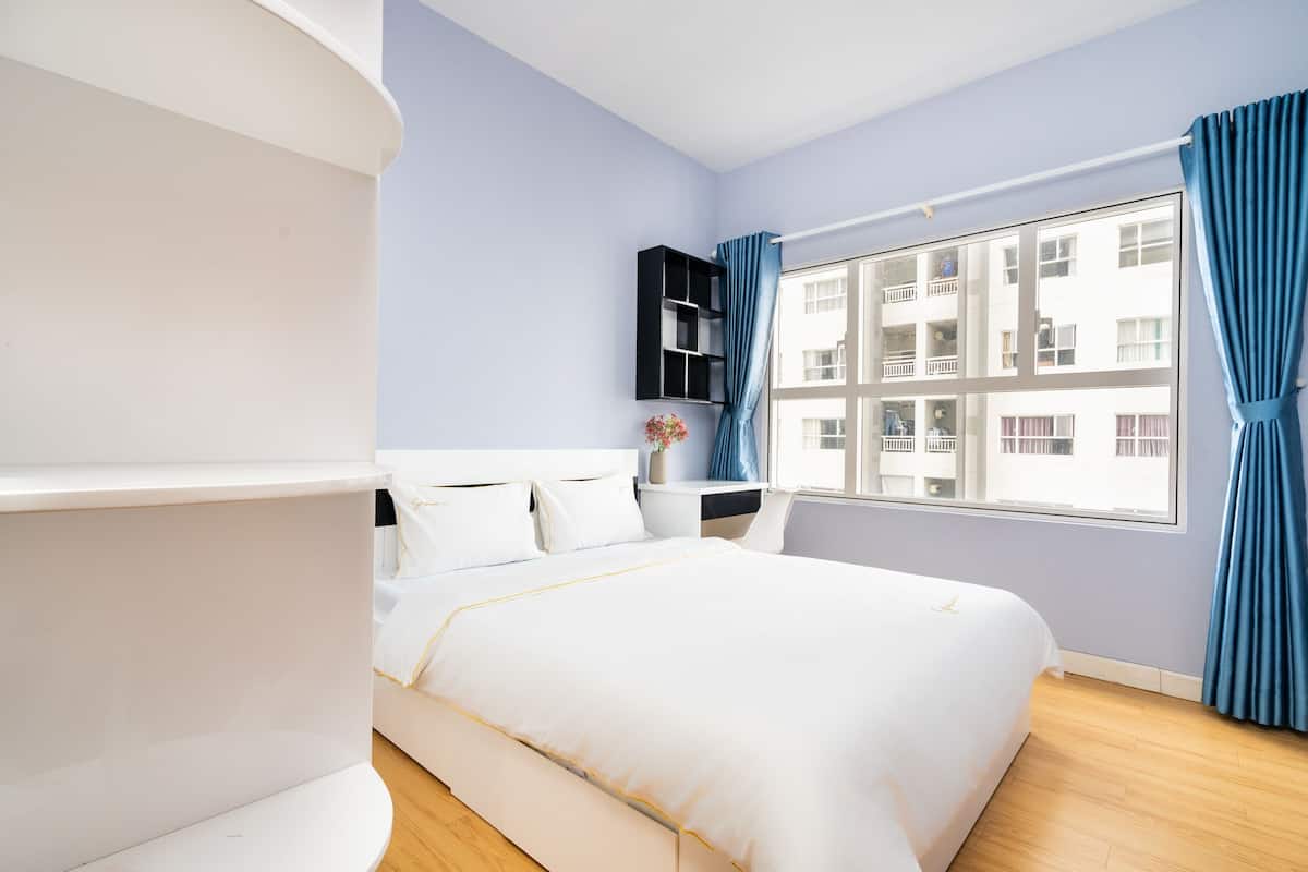 A modern bedroom with a large bed, a nightstand, a black shelving unit, and a window with blue curtains.