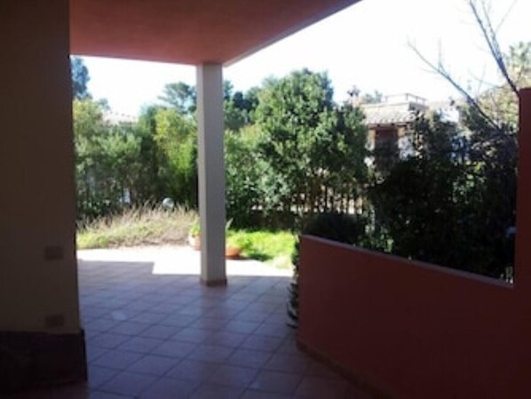 A covered patio area with tiled flooring and a view of a garden with trees and shrubs.