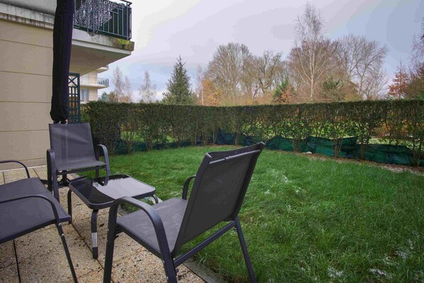 Een terras buiten met twee stoelen en een tafel, een gebouw met balkon, en een goed onderhouden haag.