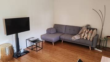Een moderne woonkamer met een grijze modulaire bank, een flatscreen televisie, een klein zwart bijzettafeltje en een decoratieve geweven kruk.