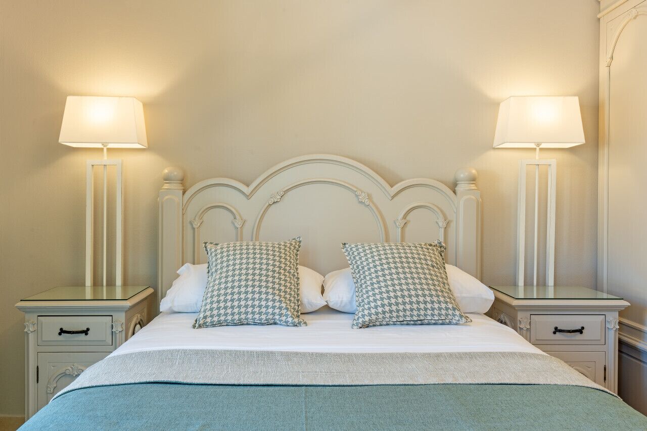 Un lit bien fait, orné de deux oreillers, avec deux tables de chevet sur lesquelles reposent des lampes, dans une pièce aux murs de couleur crème et dotée d’une tête de lit classique.