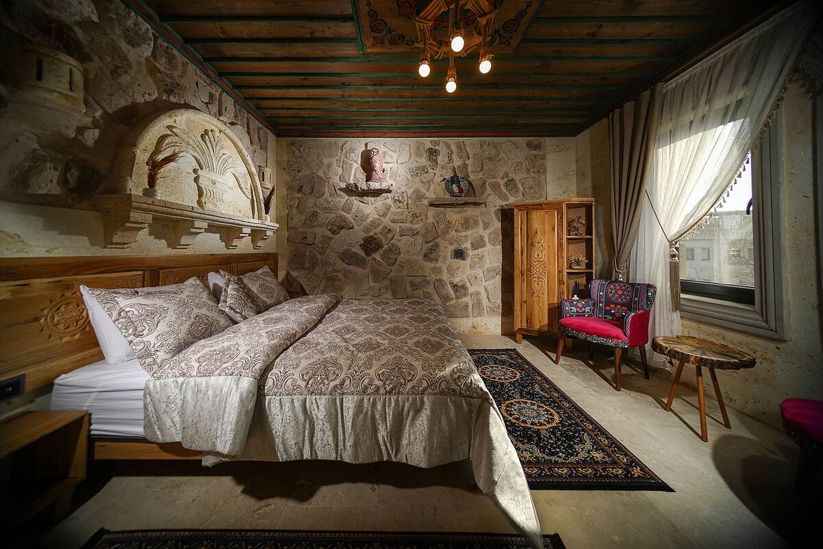 A bedroom with a stone wall, a wooden bed, a chair, a small table, and a window with curtains.
