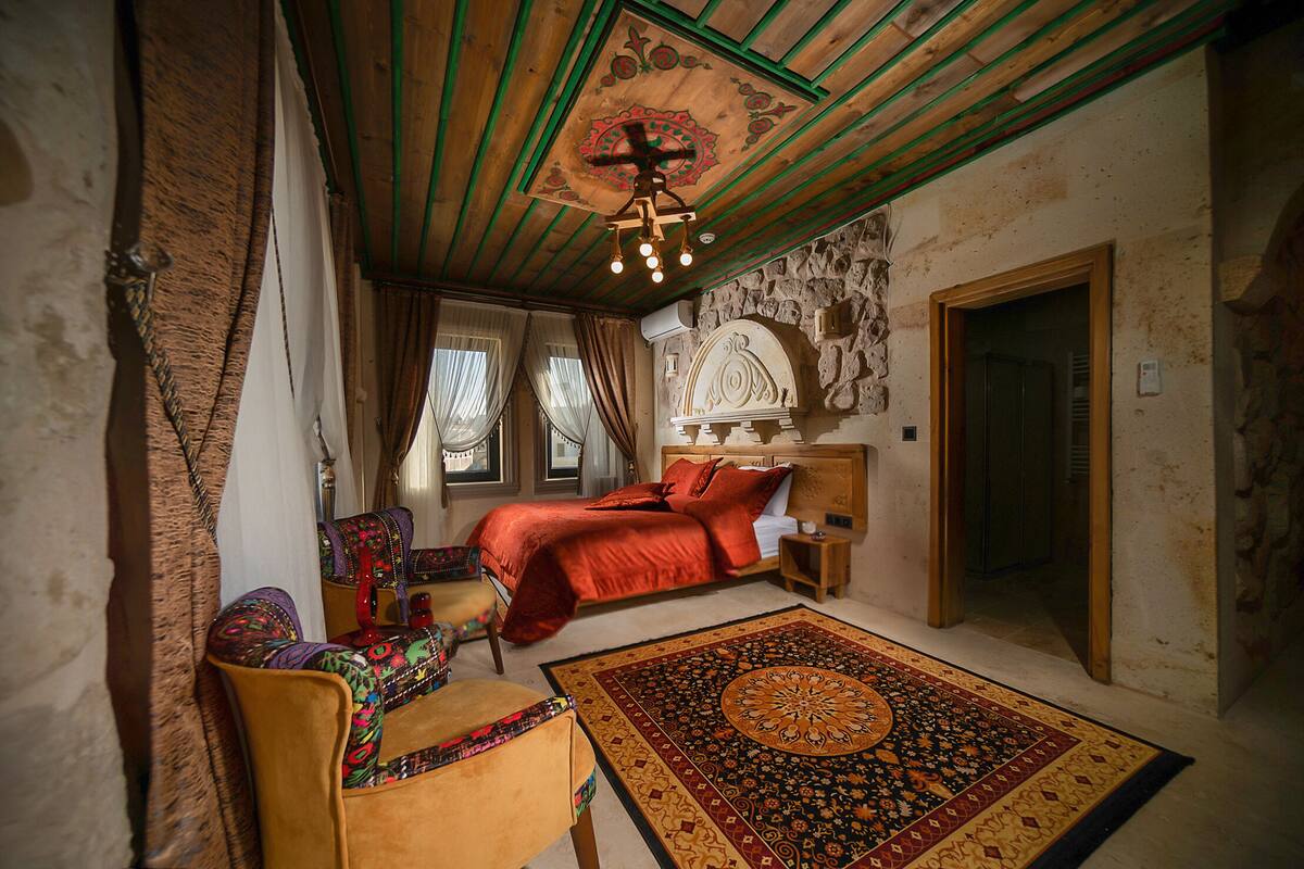 A bedroom with a wooden ceiling, a bed with red bedding, a patterned rug, and a chair with colorful upholstery.