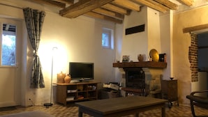 Uma sala de estar acolhedora com vigas de madeira aparentes, lareira, televisão e uma mesa de jantar.