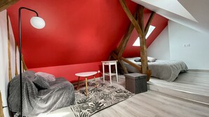 Quarto compacto e moderno com teto inclinado, parede de destaque vermelha, mesa branca pequena, puff cinza e tapete estampado.