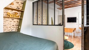 Quarto com cama, prateleira fixada na parede com a placa 'Smile', janela com persianas, televisão, mesa de madeira e uma cadeira.
