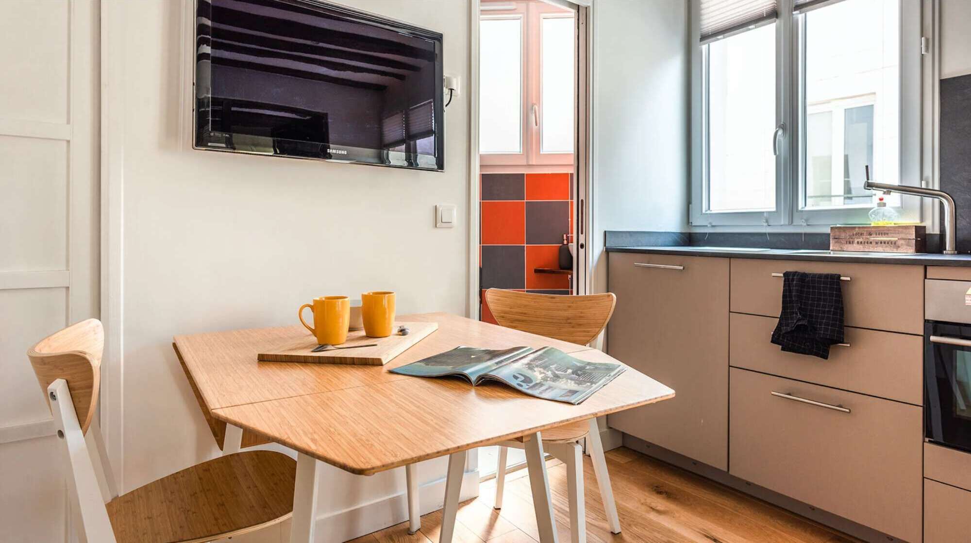 Une cuisine compacte avec un coin repas, comprenant une table en bois, des chaises, un four encastré et une fenêtre donnant sur un paysage agréable.