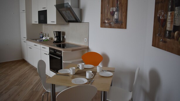 Eine moderne Küche mit weißen Schränken, einem Einbauofen und einer Kaffeemaschine. Ein Esstisch mit Stühlen und ein Gemälde von Weinflaschen sind an der Wand zu sehen.