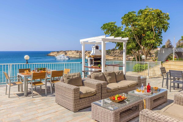 A terrace with wicker furniture, a table with fruit, and a view of the sea and boats.