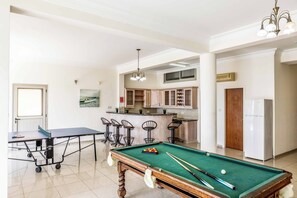 A spacious room with a pool table, a ping pong table, and a kitchen area with wooden cabinets.