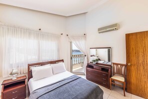 A bedroom with a bed, a dresser, a chair, a mirror, and a view of the sea.