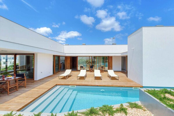 A modern outdoor pool area with lounge chairs, a wooden deck, and a clear blue sky. - Villa Ullastres Ses Alzines (Sant Lluís)