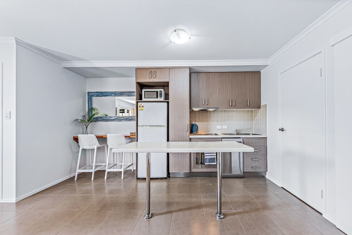 A compact kitchen with a white table, two bar stools, a microwave, a refrigerator, and wooden cabinets.