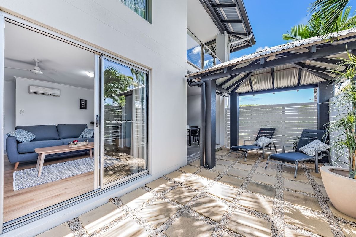 Un espace de vie moderne comprenant un canapé, une table basse et une porte-fenêtre coulissante donnant sur un patio avec des sièges d’extérieur.