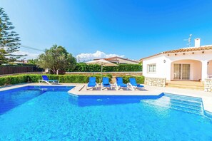 Une piscine à l’eau bleue, entourée de chaises longues et d’un bâtiment blanc doté d’un toit en tuiles.