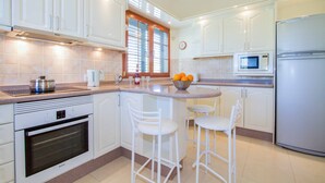 Una cocina moderna con armarios blancos, horno Bosch, microondas y refrigerador. Hay dos taburetes blancos y una ventana con persianas.