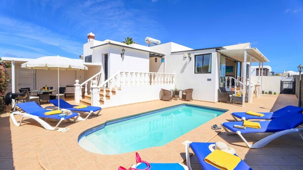 Zona de piscina con sillas reclinables azules, una piscina y un edificio blanco con una escalera cubierta.