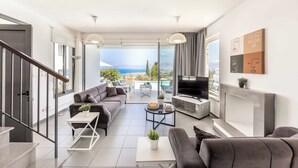 A modern living room with a sofa, coffee table, TV, and a fireplace. There is a balcony with a view of the sea. - Villa Latchi View - Three Bedroom Villa, Sleeps 6 (Neo Chorio)