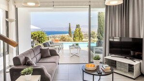 A modern living room with a sofa, TV, and a pool view. - Villa Latchi View - Three Bedroom Villa, Sleeps 6 (Neo Chorio)