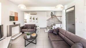 A modern living room with a sofa set, a coffee table with decorative items, and a staircase leading to an upper floor. - Villa Latchi View - Three Bedroom Villa, Sleeps 6 (Neo Chorio)