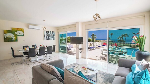 Un salon spacieux avec un grand canapé, une table basse et un coin repas comprenant des chaises et une table. Un téléviseur est fixé au mur, et un balcon donne sur une piscine et un espace extérieur aménagé avec du mobilier.