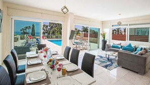 Un vaste espace de vie comprenant une table à manger pour six personnes, un canapé gris agrémenté de coussins bleus et une grande fenêtre donnant sur une piscine et des palmiers.