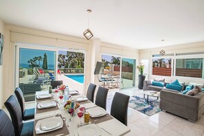 Ein geräumiger Wohnbereich mit einem Esstisch, gedeckt für sechs Personen, einem grauen Sofa mit blauen Kissen und einem großen Fenster mit Blick auf einen Pool und Palmen.