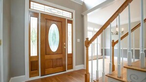Una puerta principal de madera con un panel de vidrio, una escalera con balaustres blancos y una lámpara sobre ella.