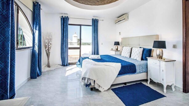 A bedroom with a large bed, a white nightstand, a blue rug, and a view of a white building through the window. - Villa Gira - Four Bedroom Villa, Sleeps 8 (Albufeira)