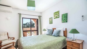 Ein Schlafzimmer mit einem Bett, einem Nachttisch, einem Sessel und Blick ins Freie durch eine Schiebetür.
