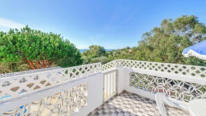 Un balcone con ringhiera bianca a traliccio, pavimento piastrellato e vista su alberi e mare.