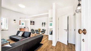 Ein modernes Wohnzimmer mit einer schwarzen Couch, einem Esstisch und Stühlen. Im Hintergrund befindet sich ein Küchenbereich mit Schränken und einer Arbeitsplatte.