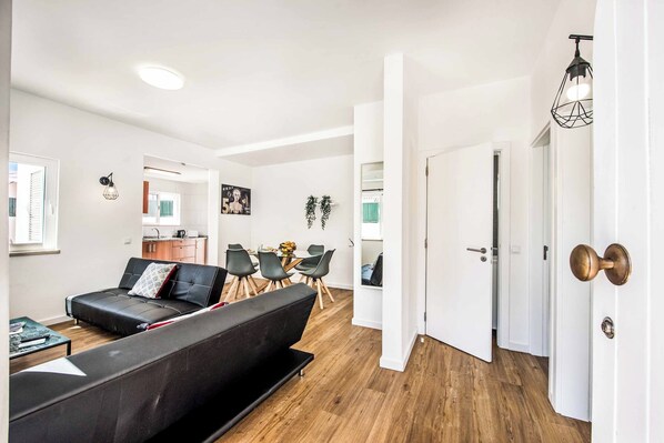 Un salon moderne avec un canapé noir, une table à manger et des chaises. On aperçoit, au fond, un coin cuisine avec des placards et un plan de travail.