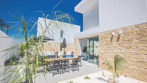 A modern outdoor patio with a dining table and chairs, surrounded by stone walls and palm plants. - Vie Bleu 1 - Four Bedroom Villa, Sleeps 8 (Πρωταράς)